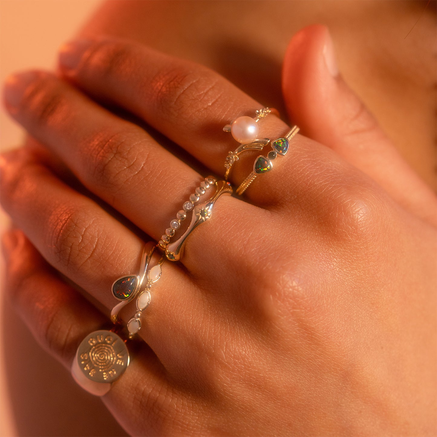 A close-up of a hand wearing the Solid Gold Black Opal Galaxy Droplet Ring, surrounded by other gold rings with pearls, gemstones, and an engraved round pendant, set against a warm-toned background.
