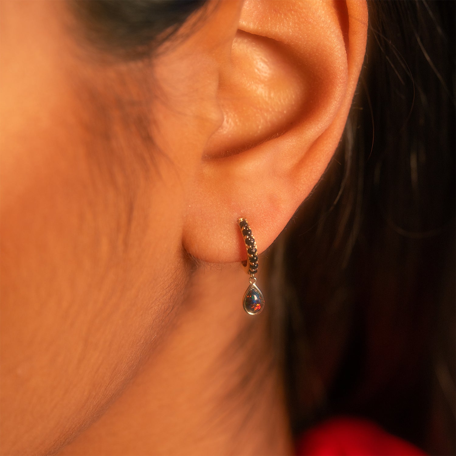 Close-up of a person's ear wearing the Solid Gold Black Opal Galaxy Droplet Huggie Hoops, featuring a teardrop-shaped black opal pendant. A softly blurred background accentuates the earring's intricate detail.