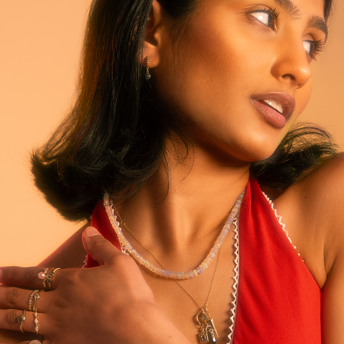 A woman in a red dress with lace trim poses against a warm beige background, wearing layered necklaces and a Solid Gold Black Opal Divine Direction Ring, her hand resting near her neckline.