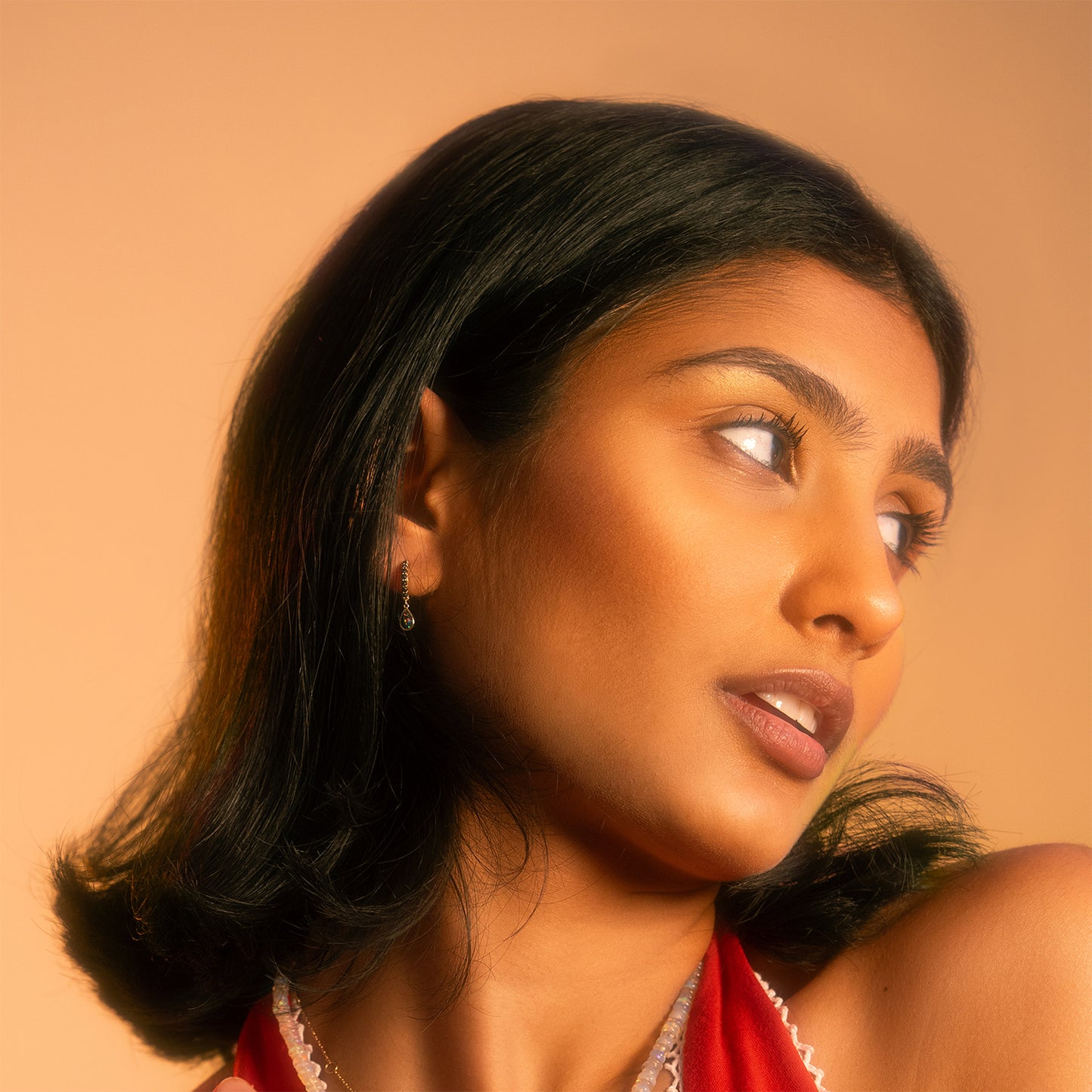 A woman with medium skin and shoulder-length black hair looks to the side against a peach background. She wears a red top, natural makeup, and Solid Gold Black Opal Galaxy Droplet Huggie Hoops under soft lighting highlighting her features.