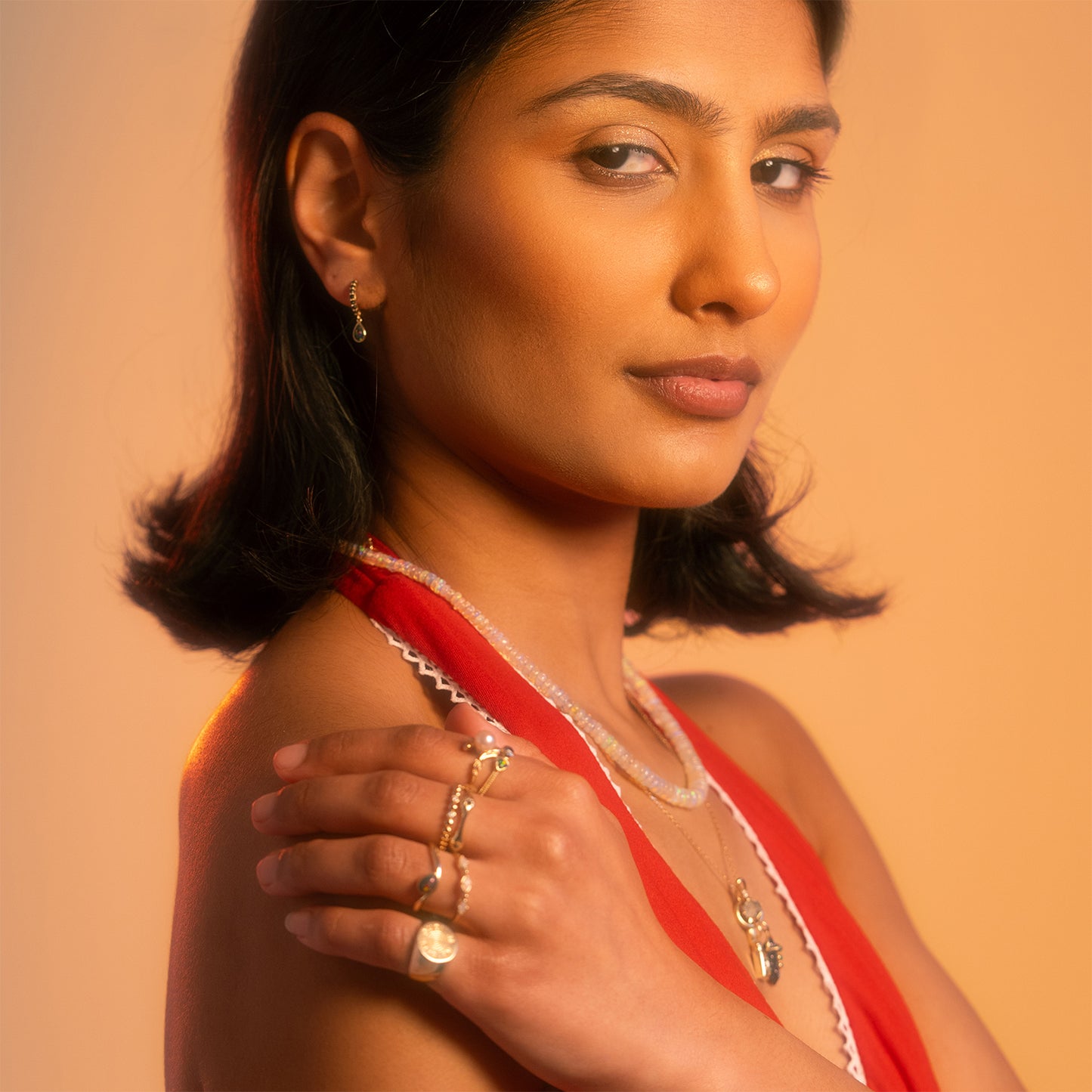 A woman with short dark hair and a red top showcases the Solid Gold Black Opal Galaxy Droplet Huggie Hoops, posing with her hand on her shoulder against a warm, softly lit background.