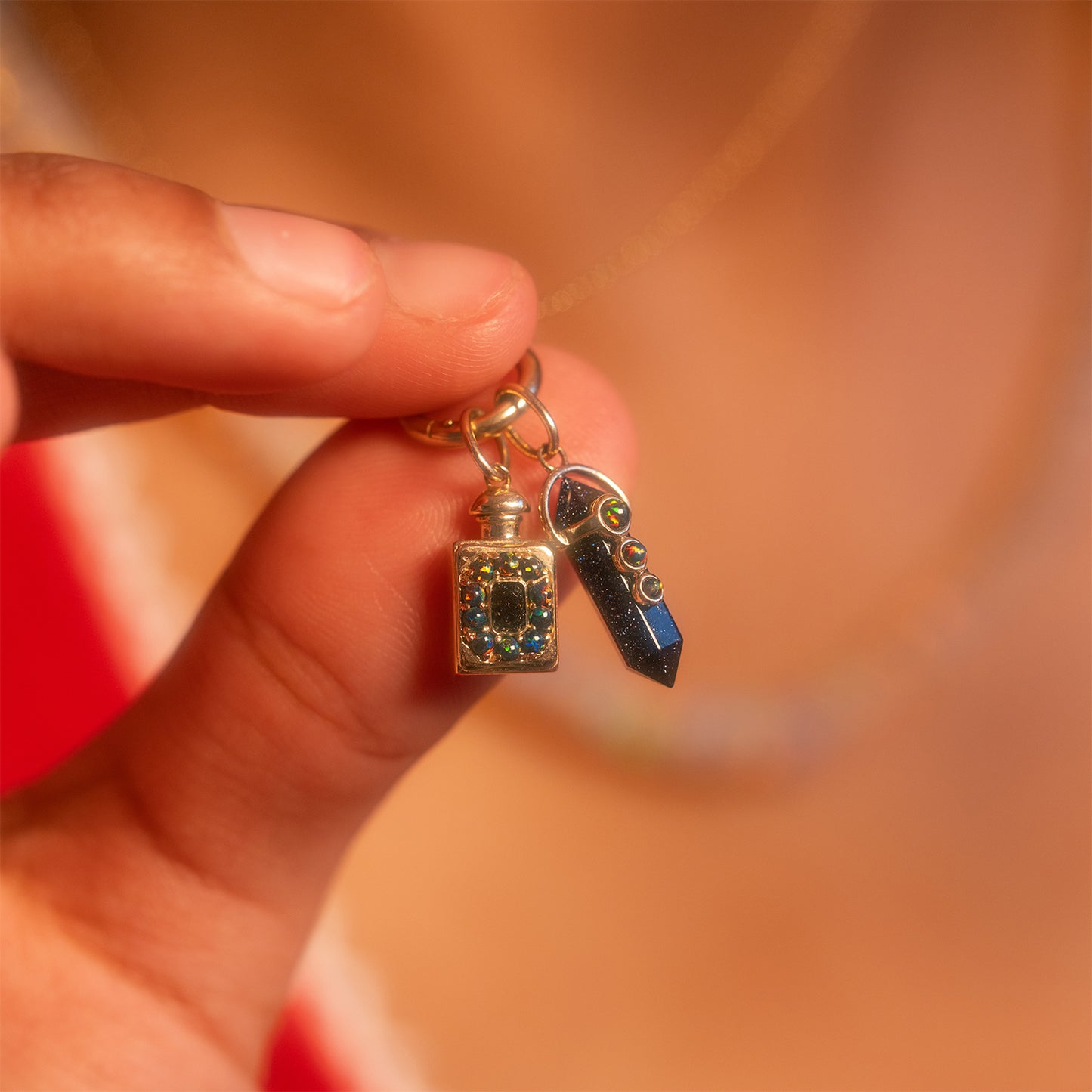 A hand holds a Local Eclectic exclusive Solid Gold Amulet Charm keychain featuring two charms—a glittery bottle and a black crystal-shaped pendant—set against a softly blurred background.