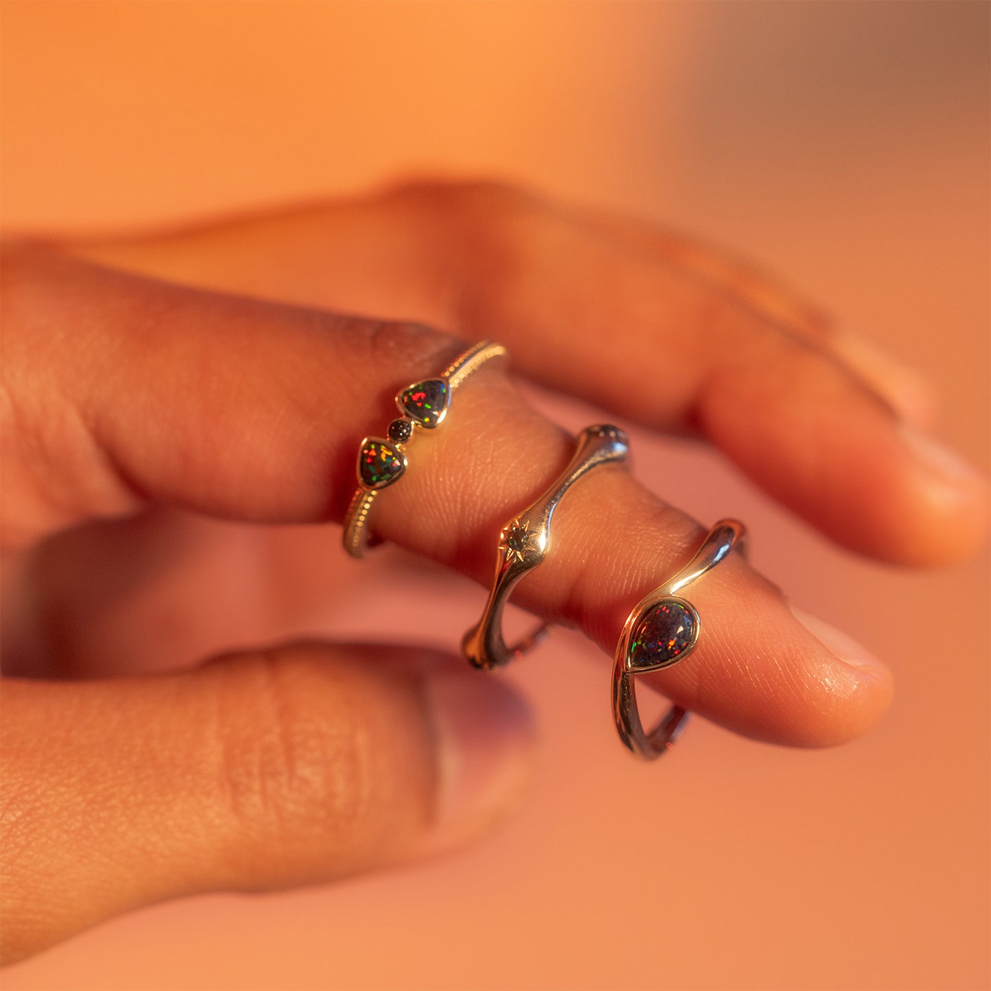 A close-up of a hand wearing three Solid Gold Black Opal Galaxy Droplet Rings, each featuring small, round, multicolored opals, set against a warm, softly blurred background.