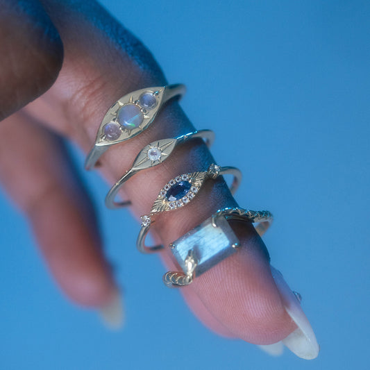 A close-up of a hand wearing four gold rings, including the Solid Gold Iolite Evil Eye Ring, each with unique designs, showcased on the index finger against a soft blue background.