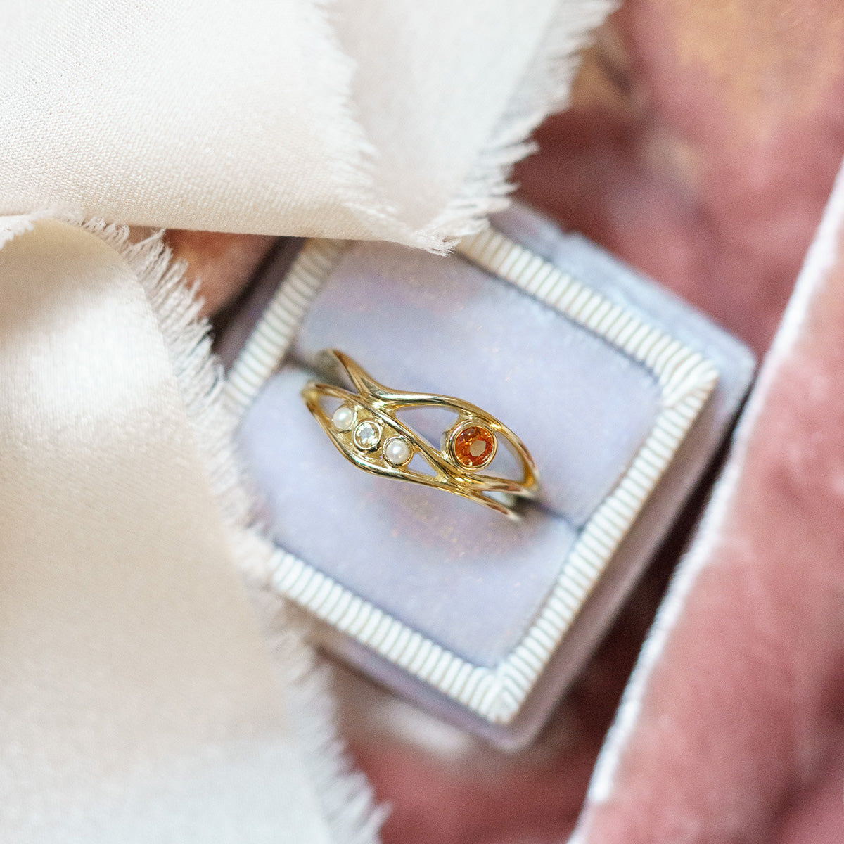 The 10k Orange Sapphire and Citrine Bayside Ring features three clear stones and one orange stone, showcased in a light purple ring box with soft pale pink and cream fabric.
