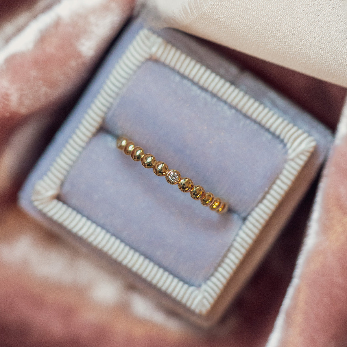 The 10k Hidden Message Morse Code Diamond Band features a delicate gold design with a central diamond, shown in a light purple velvet ring box trimmed in cream.
