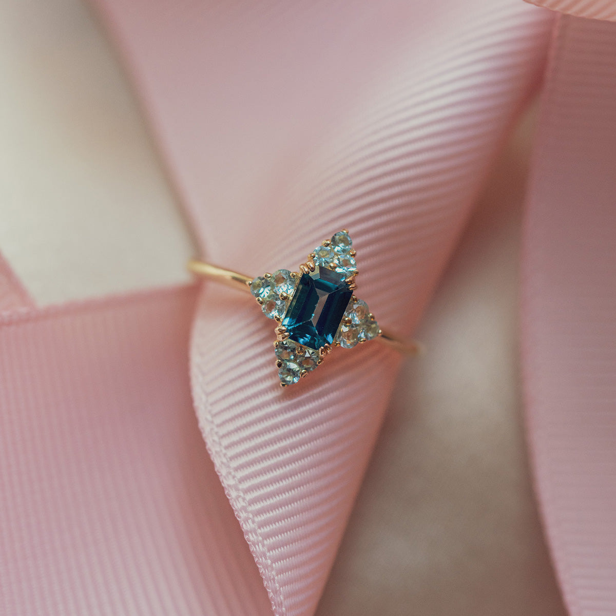 The 10k Snow Queen Blue Topaz Cluster Ring features a rectangular blue topaz at the center, encircled by clusters of clear stones, and is displayed on folded pink fabric.
