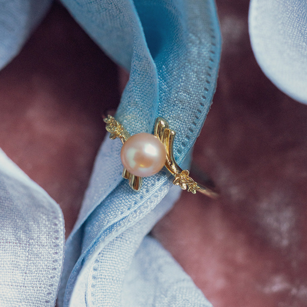 The 10k Forget Me Not Pink Pearl Bypass Ring, featuring a central pink pearl and diamond accents, is displayed on soft light blue fabric against a blurred mauve background.