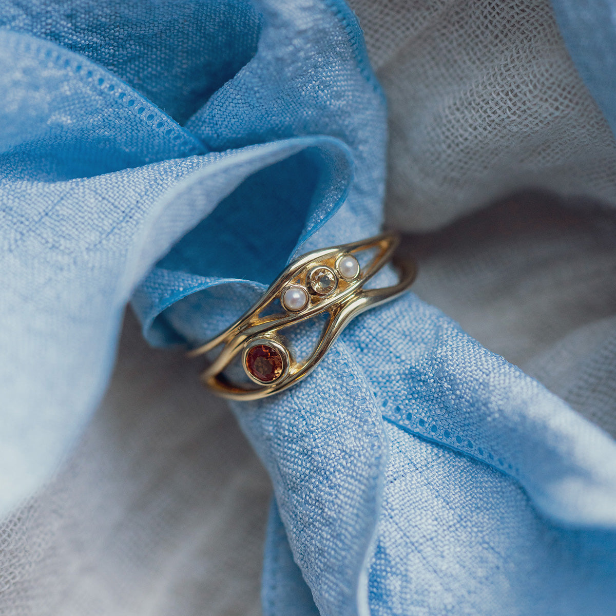 A 10k yellow gold Bayside ring with an orange sapphire and citrine rests on soft, textured blue and white fabric.