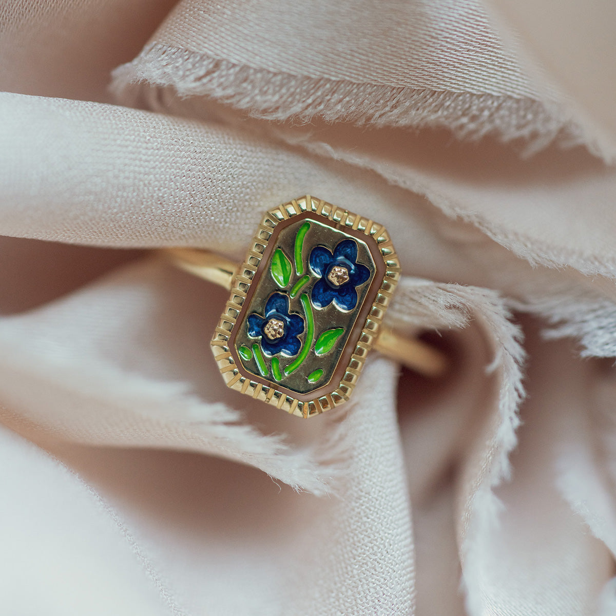 The 10k Gallery Frames Enamel and Malachite Reversible Ring features a rectangular face with two blue enamel flowers, green leaves, and diamond centers, displayed on soft light beige fabric.