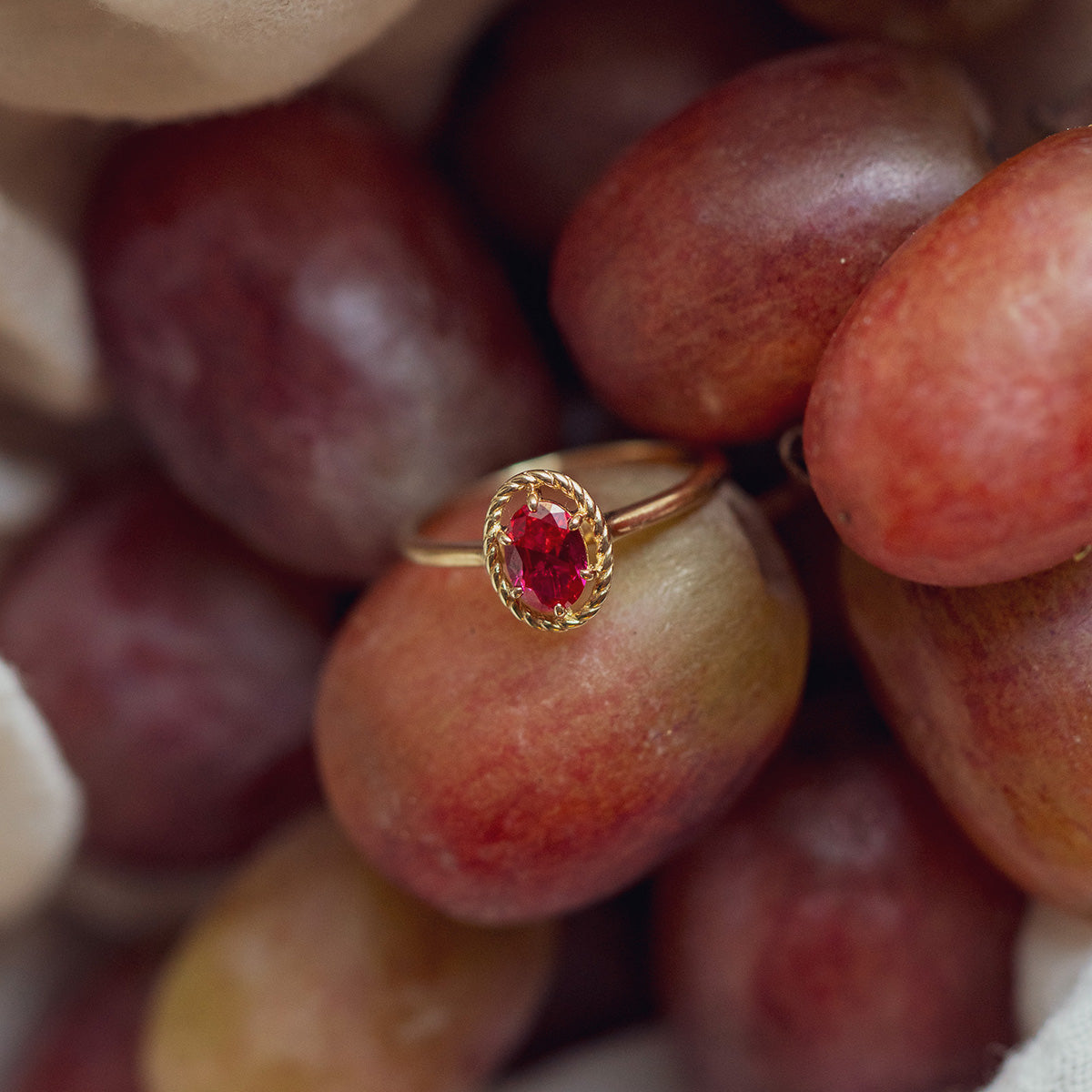 The 10k Lucky In Red Ruby Halo Ring, featuring an oval red ruby, is showcased atop a cluster of red and green grapes.