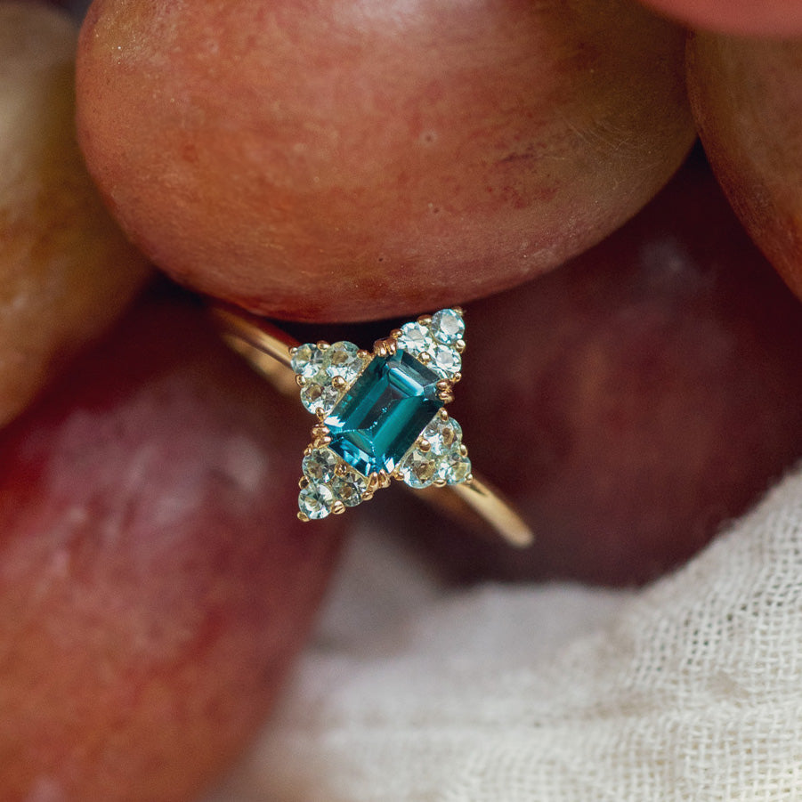 The 10k Snow Queen Blue Topaz Cluster Ring, featuring a rectangular teal gemstone surrounded by small white diamonds in a cross pattern, is displayed against a backdrop of reddish grapes.