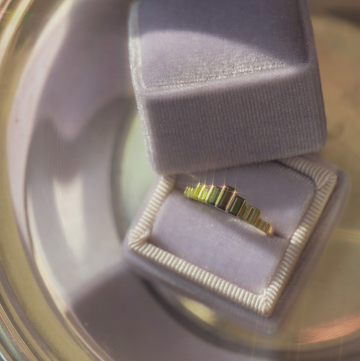 The 10k Green Tourmaline Skyline Band, featuring rectangular green gemstones, is showcased in a light purple velvet ring box as soft sunlight casts gentle reflections and shadows.
