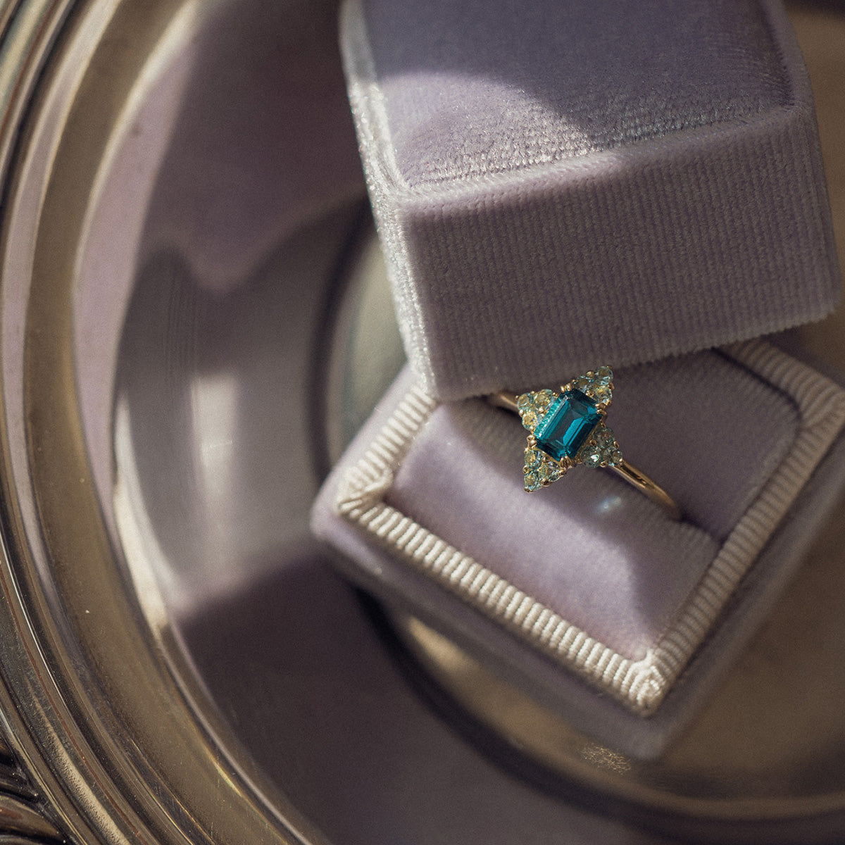 The 10k Snow Queen Blue Topaz Cluster Ring, featuring a blue rectangular gemstone and diamond accents, is displayed in an open light purple velvet ring box on a reflective surface.