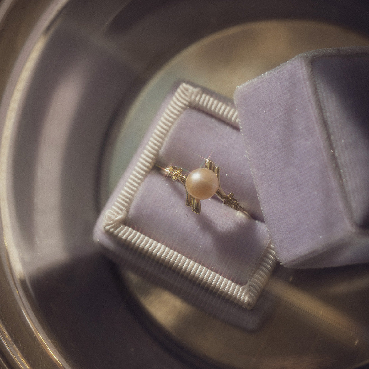 The 10k Forget Me Not Pink Pearl Bypass Ring, featuring a central pink pearl and diamond accents, is displayed in a light purple velvet ring box next to another similar box on a reflective surface.