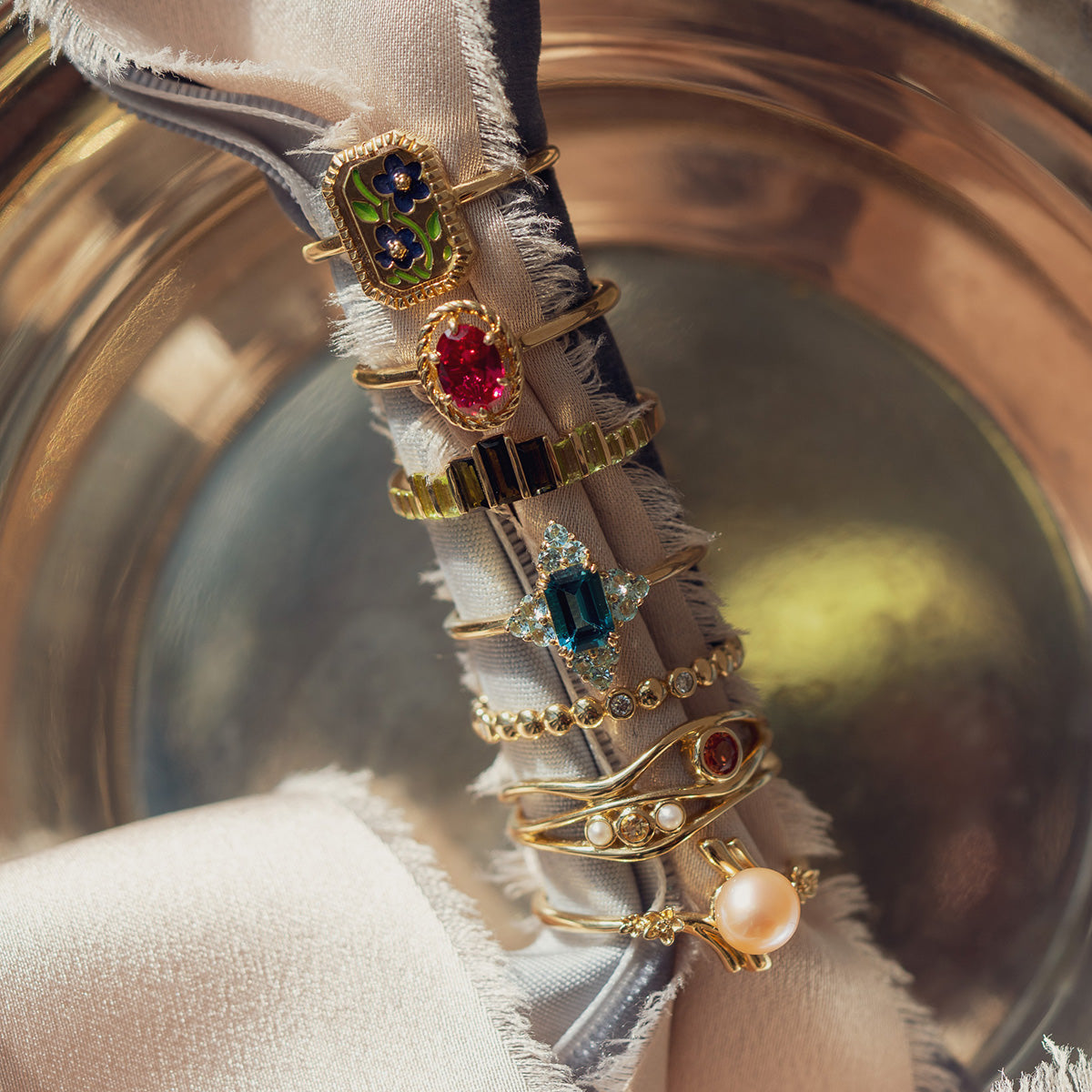 The 10k Green Tourmaline Skyline Band, featuring a vivid gemstone set in gold, is displayed elegantly on textured fabric with a gleaming metallic backdrop.