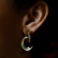 Close-up of an ear with three earrings: a small silver cartilage stud, a gold star-shaped stud, and La Lune Teal Green Earrings—a gold crescent moon dangle with a green tint and gemstone detail.