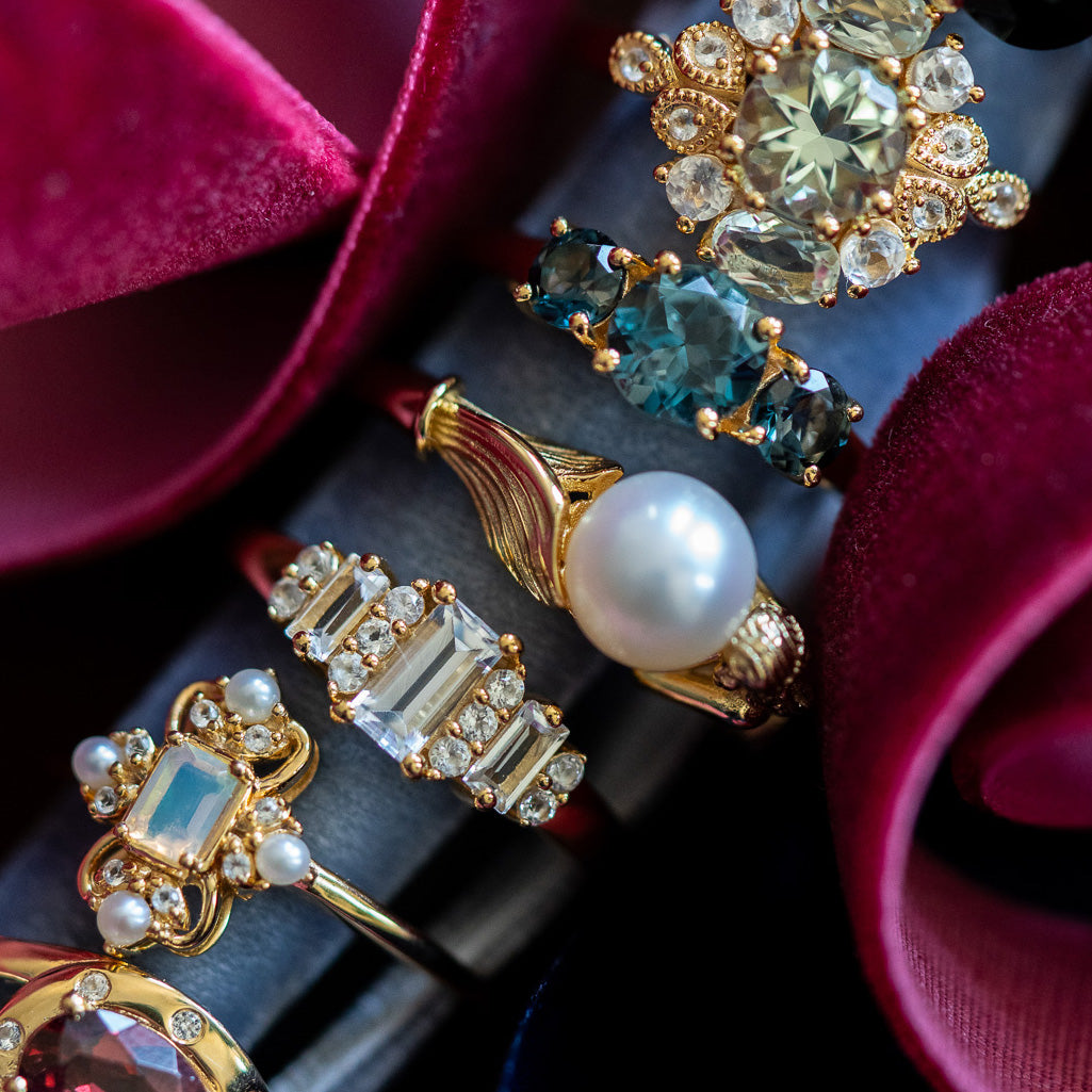 A close-up of five ornate 14k Gold Vermeil rings, featuring gemstones and pearls—including the Royal Gardens Green Amethyst Cocktail Ring—shown against lush red velvet with blue, clear, and white stones in intricate designs.