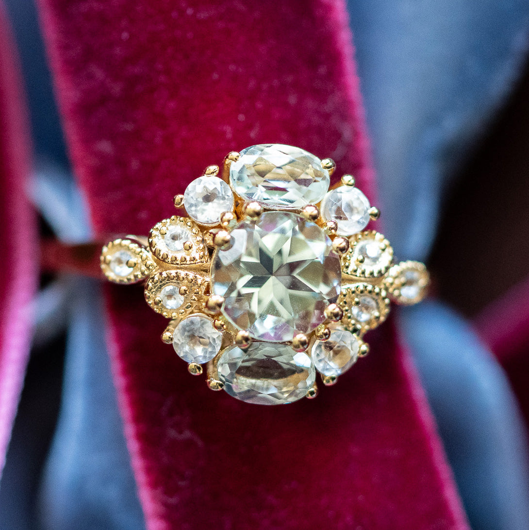 A close-up of the Royal Gardens Green Amethyst Cocktail Ring, crafted in 14k Gold Vermeil with a central oval green amethyst and smaller clear stones, displayed on deep pink velvet with blurred blue fabric in the background.