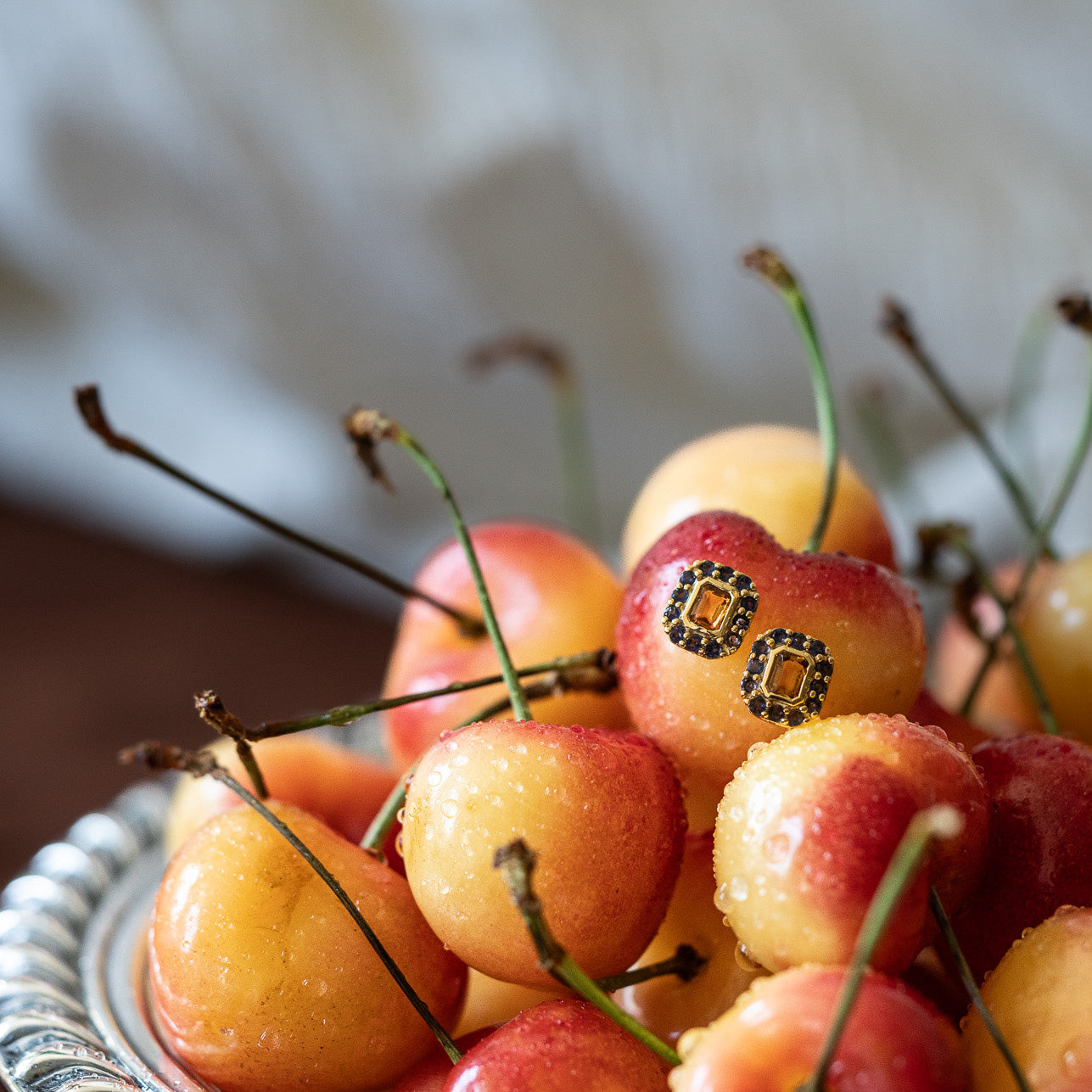A bowl of fresh Rainier cherries features one cherry with playful cartoon eyes, ready to accessorize with Citrine and Iolite Novella Studs for extra charm.