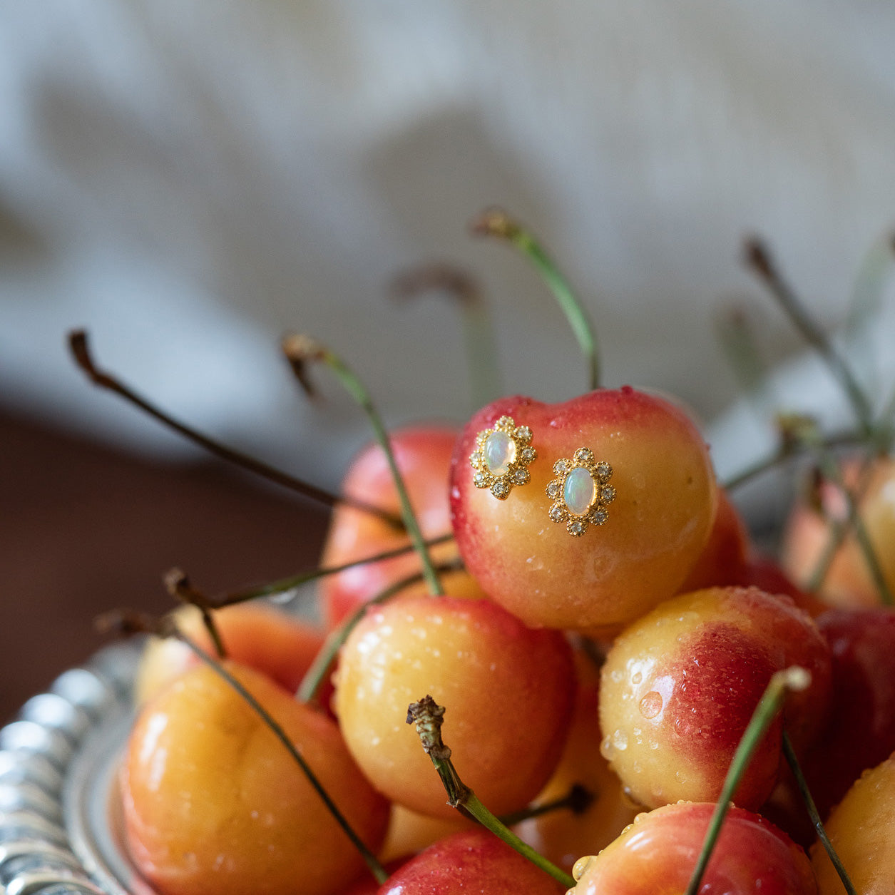 A close-up of Rainier cherries in a silver bowl features one cherry adorned with Opal Majesty Studs, adding a whimsical touch. The cherries are fresh and glistening with water droplets.