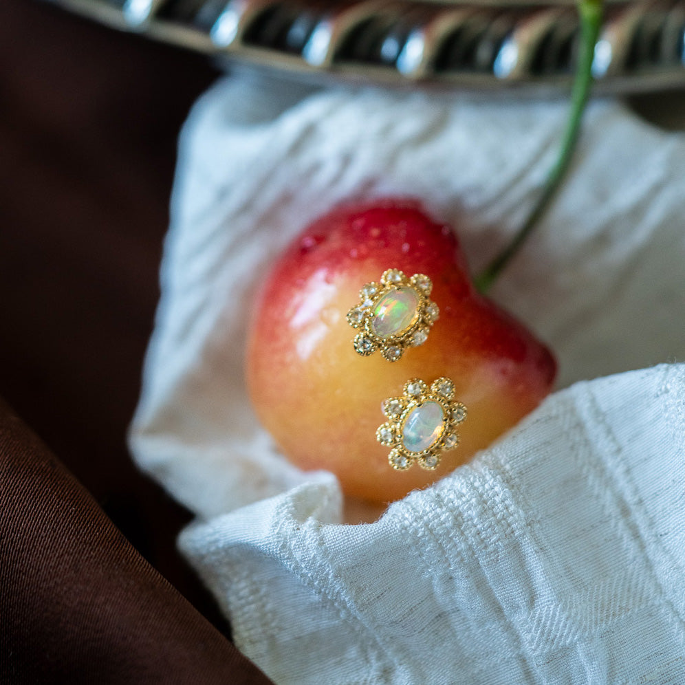 The Opal Majesty Studs, featuring a vintage-inspired floral design, are displayed on a red and yellow cherry atop white textured cloth, with a silver tray and brown fabric in the background.