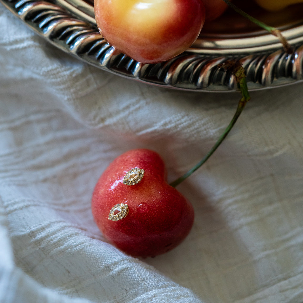 A close-up of a yellow-red cherry with two small damaged spots on its surface, lying on textured white cloth near a silver dish and sparkling Green Amethyst Marquise Muse Studs.