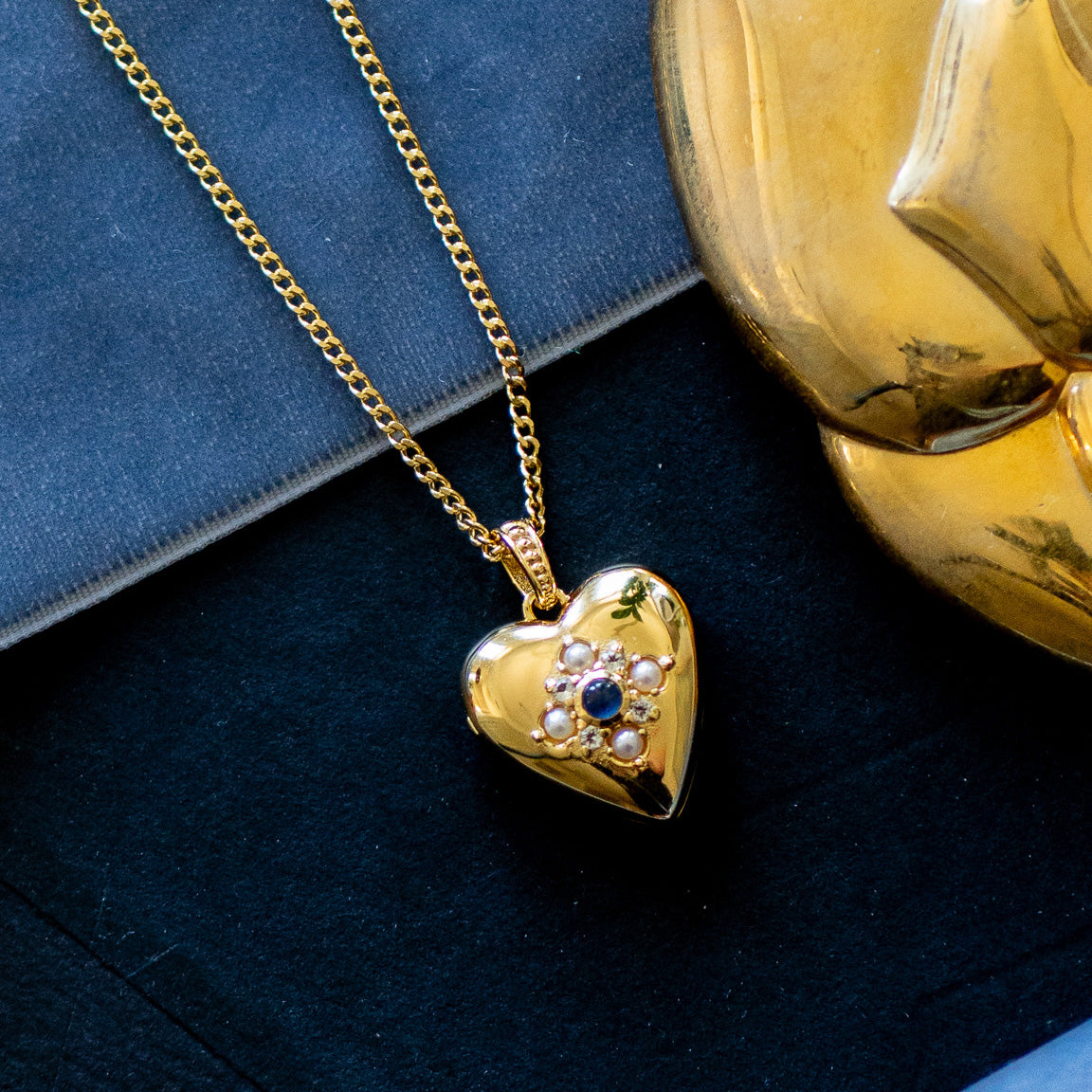 Rose’s Blue Sapphire Heart Locket, a gold vermeil heart-shaped pendant with blue sapphire and floral details, hangs on a gold chain and is displayed on a dark surface beside a gold object and a dark book.