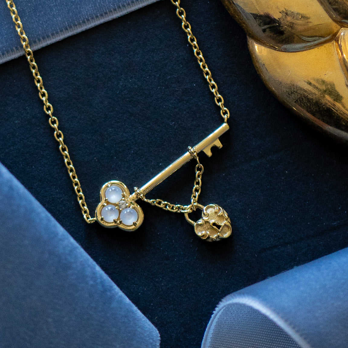 The Lock and Key Necklace features a gold vermeil skeleton key pendant with three white stones and a small heart charm, shown against a dark background with blue and gold accents.
