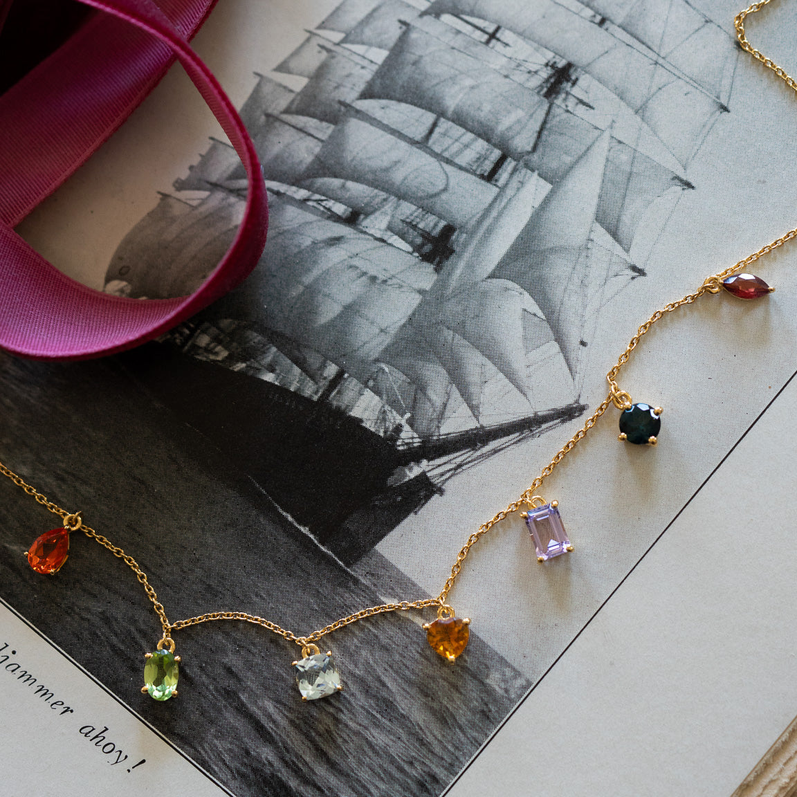 The Classical Confetti Gemstone Necklace, featuring delicate 14k gold vermeil and colorful gemstone pendants, is displayed on a black-and-white sailing ship photo beside a pink ribbon—a beautiful vintage-inspired jewelry piece.