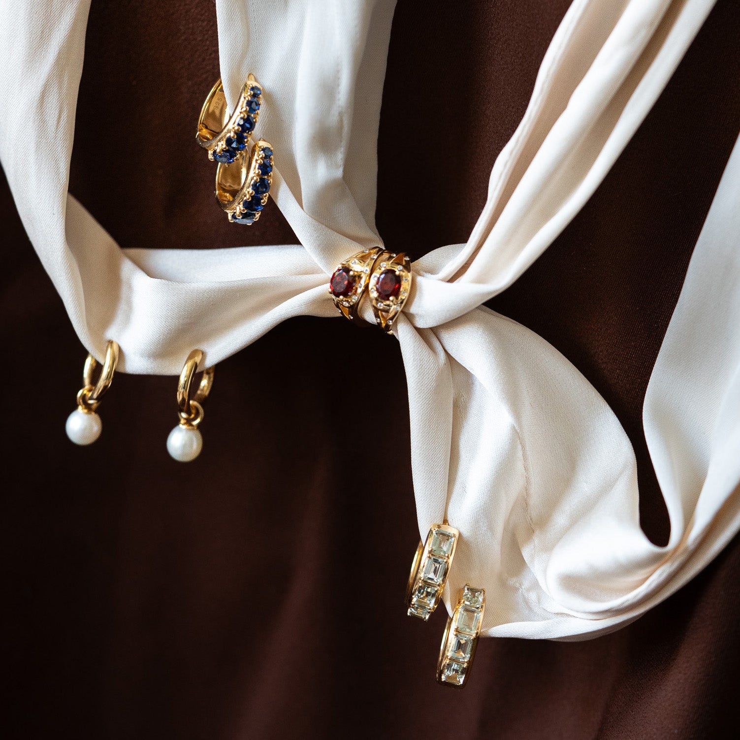 The Gilded Garnet Huggie Hoops—gold vermeil earrings with pearls, garnets, white topaz, and blue stones—hang from a white knotted fabric set against a brown background.