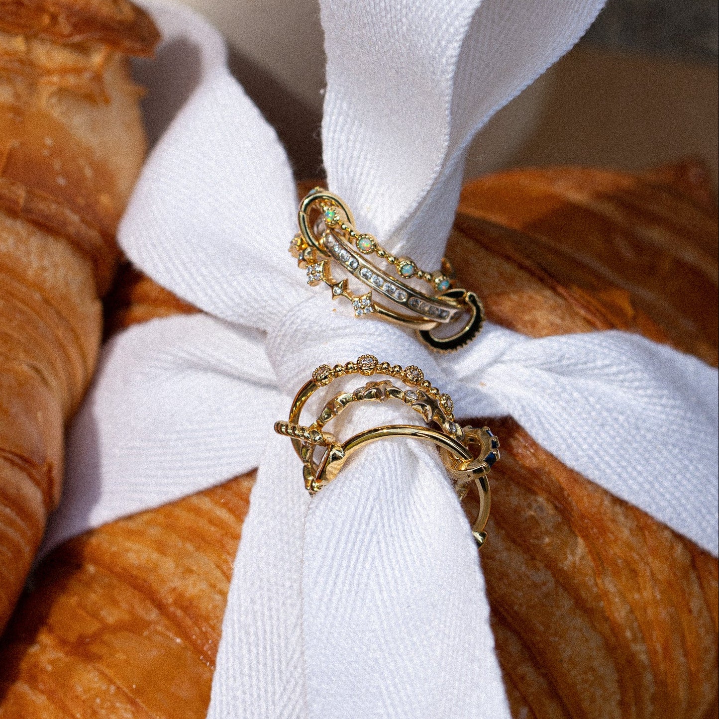 The Bold Eternity Stacking Ring, featuring an intricate design with sparkling gems, is displayed on a white ribbon tied in a bow, with croissants visible in the background.