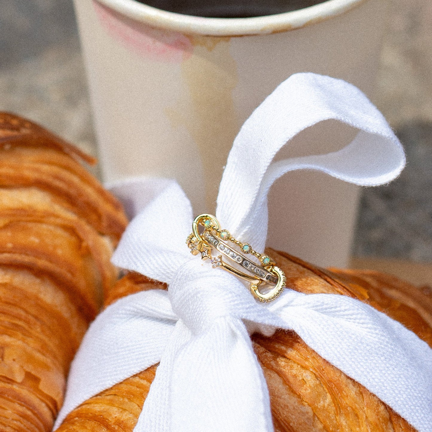 A close-up of a croissant tied with a white ribbon adorned by the Starlet Infinity Stacking Ring, set in front of a white paper coffee cup.