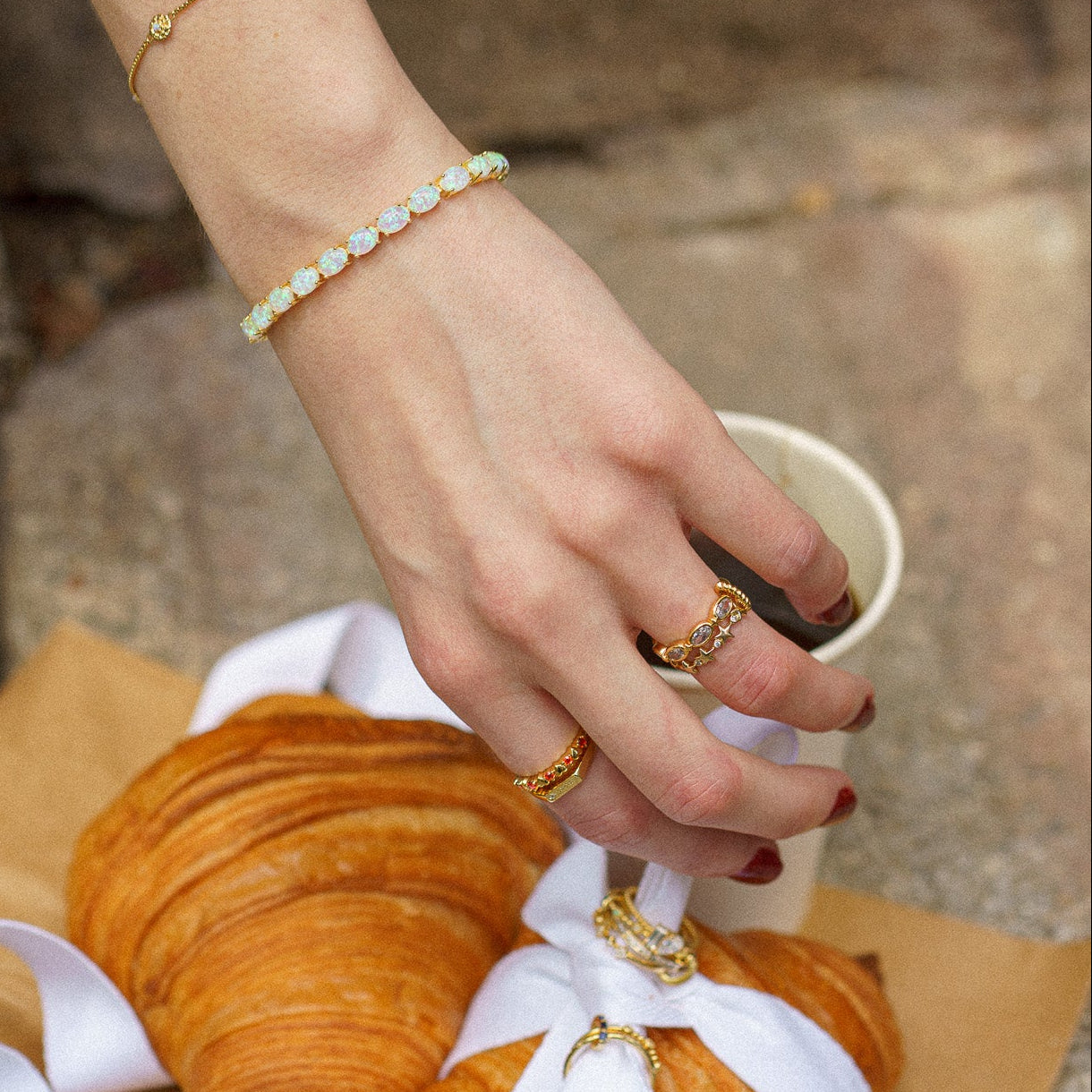 A hand with red nail polish wears the Bold Moon Beam Stacking Ring and a gemstone bracelet, reaching over croissants wrapped in a white ribbon and a cup, all set on a rustic surface.