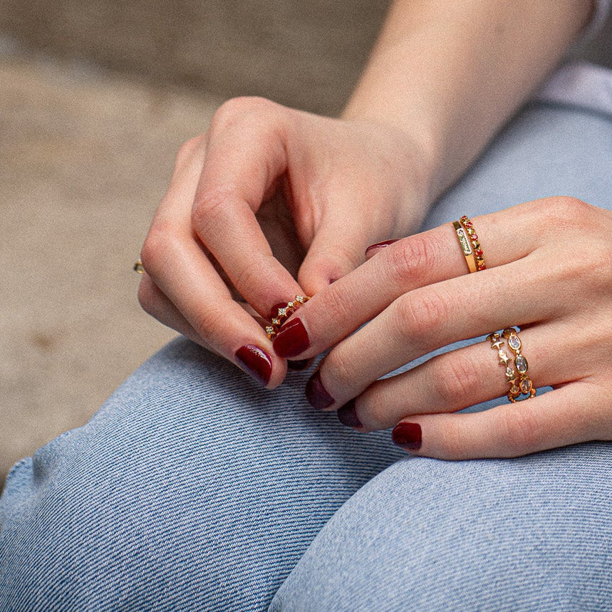 Close-up of hands with dark red nails, wearing the Starlet Infinity Stacking Ring and other gold gemstone rings, gently touching over light blue denim jeans. The background is softly blurred.