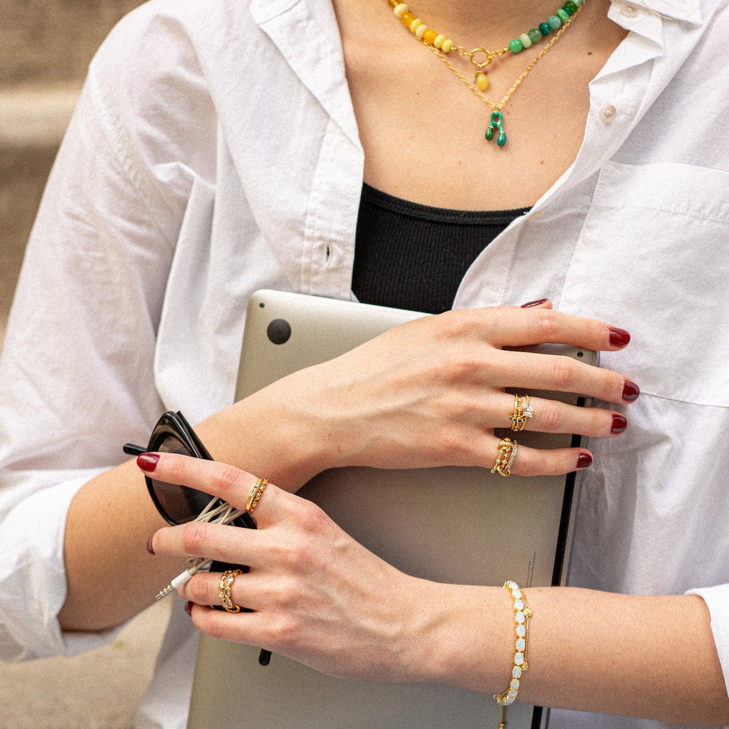 A person in a white shirt and black top holds a tablet and sunglasses, accessorized with the Twisted Toi Et Moi Ring, gold bracelets, layered beaded necklaces, and dark red nails.