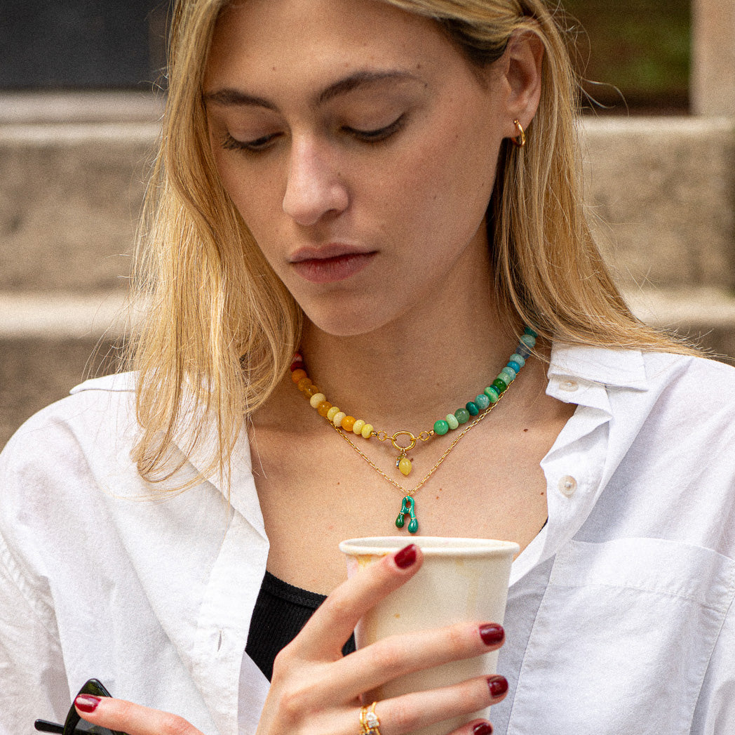 A woman with long blonde hair, in a white shirt and gold rings and earrings, wears the Beaded Gemstone Charm Holder Necklace. She has red nail polish, holds a white paper cup, and looks down thoughtfully.