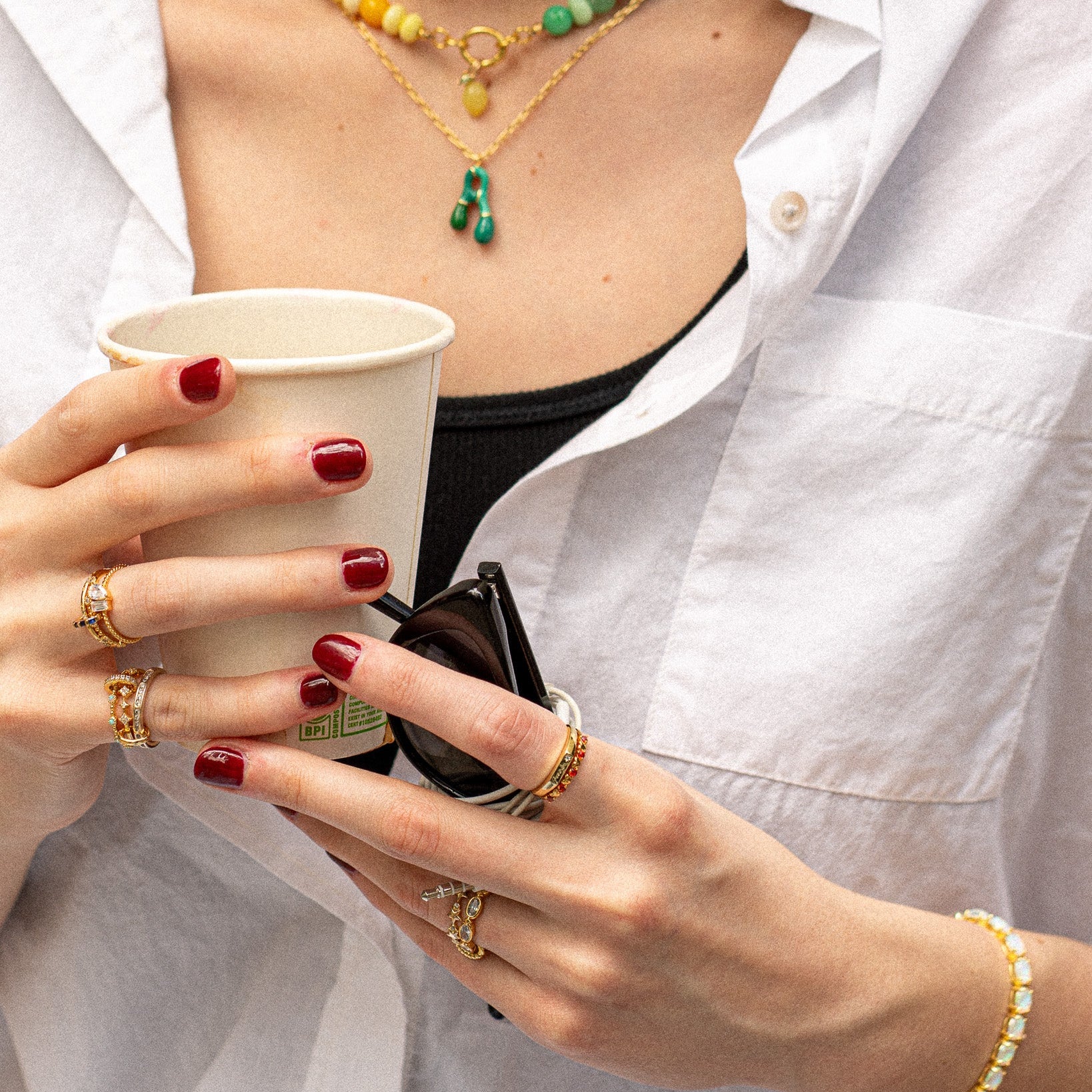 A person with red painted nails, wearing the Starlet Infinity Stacking Ring along with other rings and bracelets, holds a paper coffee cup and sunglasses. They have on a white shirt and layered necklaces; only their hands and torso are shown.