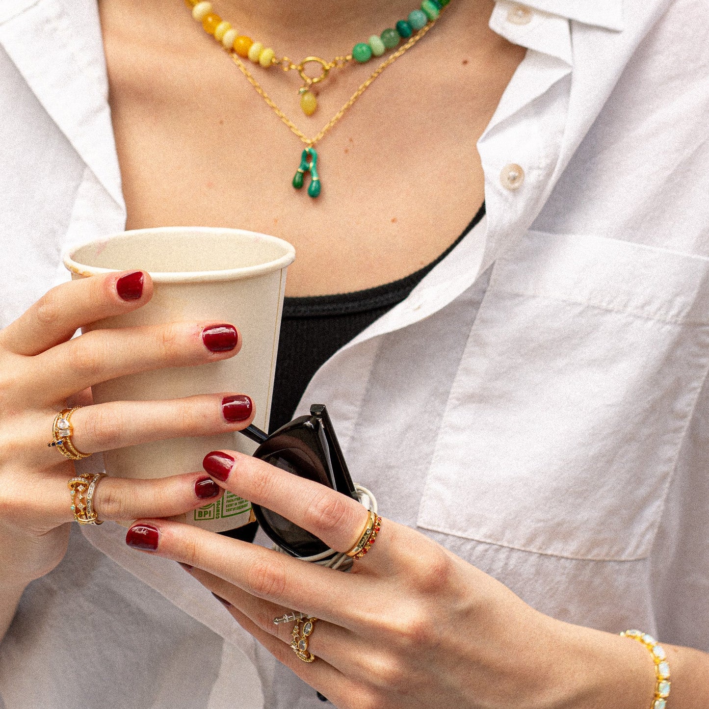A person with dark red nails wears several gold rings, including the Dainty Flicker Stacking Ring, plus bracelets and layered necklaces. They hold a paper cup and sunglasses while wearing a white shirt.