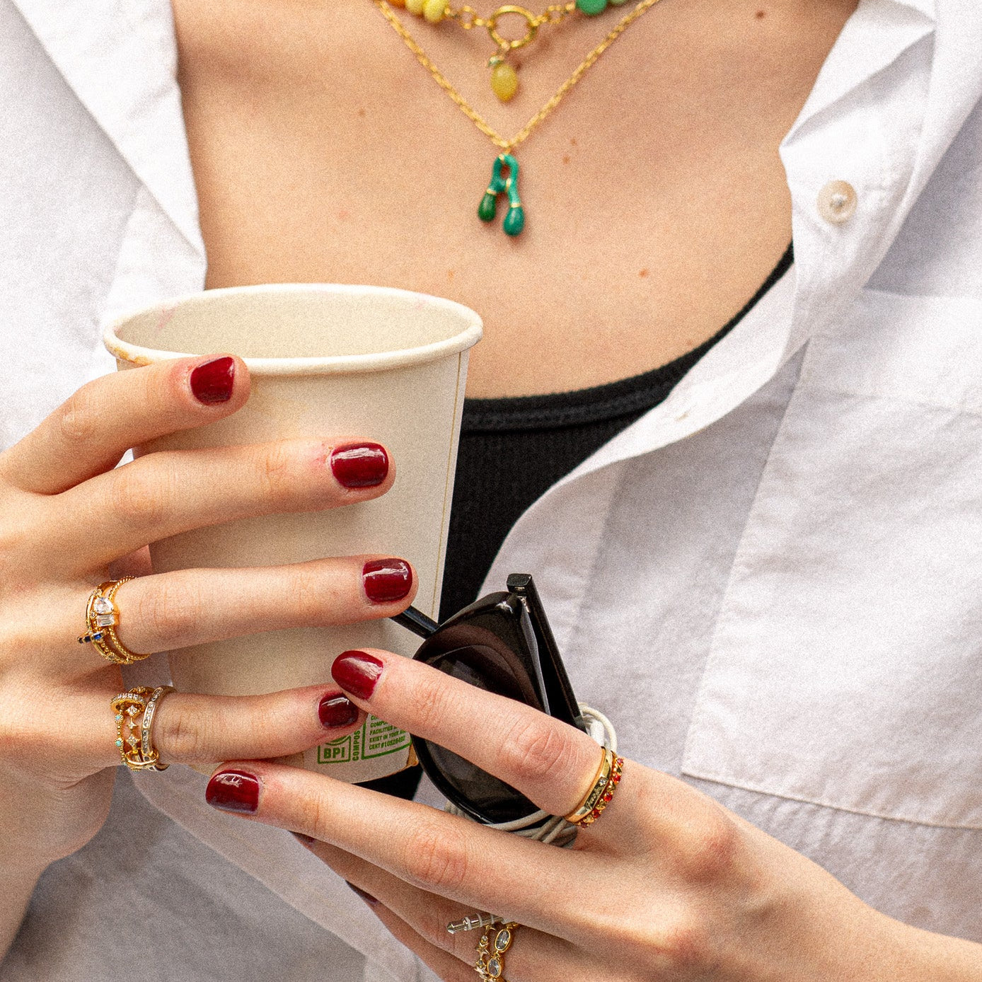 A person wearing a Twisted Toi Et Moi Ring, red nail polish, and gold necklaces with colorful pendants holds a paper cup and sunglasses while dressed in a white shirt and black top.