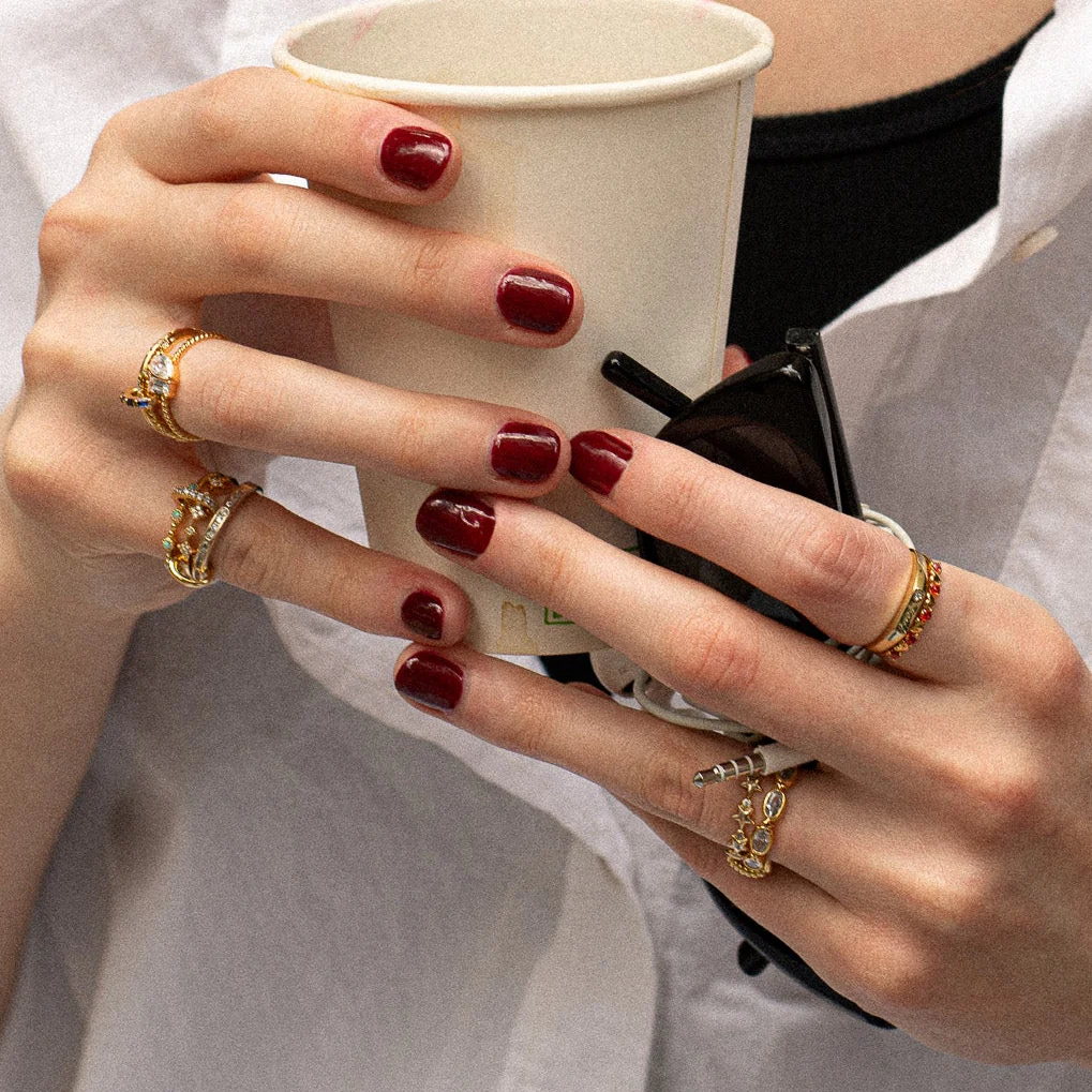 Close-up of hands with dark red nail polish wearing the Bold Moon Beam Stacking Ring, holding a white paper cup and black sunglasses. The person is dressed in a white shirt and black top.