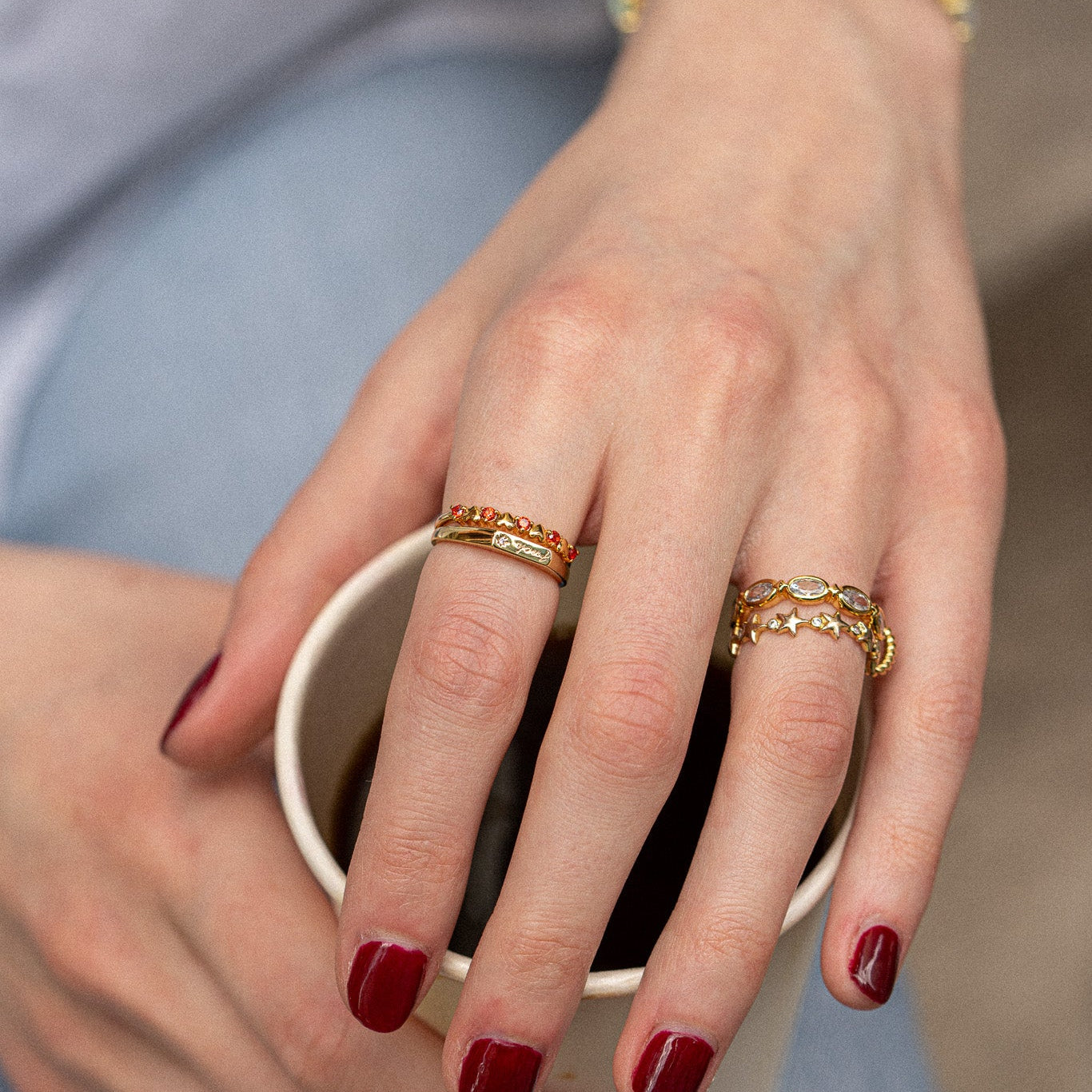 A close-up of a hand with dark red nail polish holding a white cup, wearing the Starlet Infinity Stacking Ring and two other gold rings with gemstone and star designs. The person is dressed in light blue jeans.