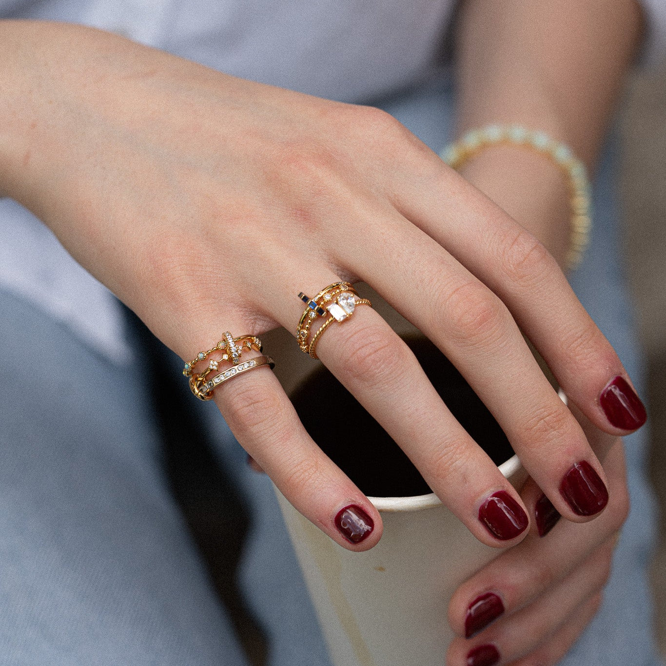 A hand with dark red nails holds a paper cup. The person wears the Bold Eternity Stacking Ring with other gold rings and a beaded bracelet, paired with light blue denim.