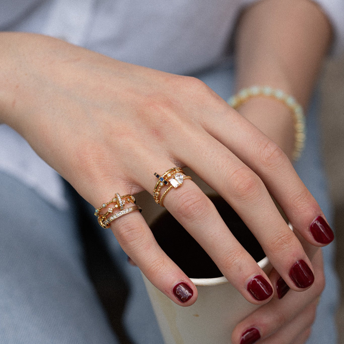 A hand with dark red nails holds a cup, adorned with the Dainty Flicker Stacking Ring and gemstone rings, along with a beaded bracelet. The wearer is dressed in a light-colored shirt.