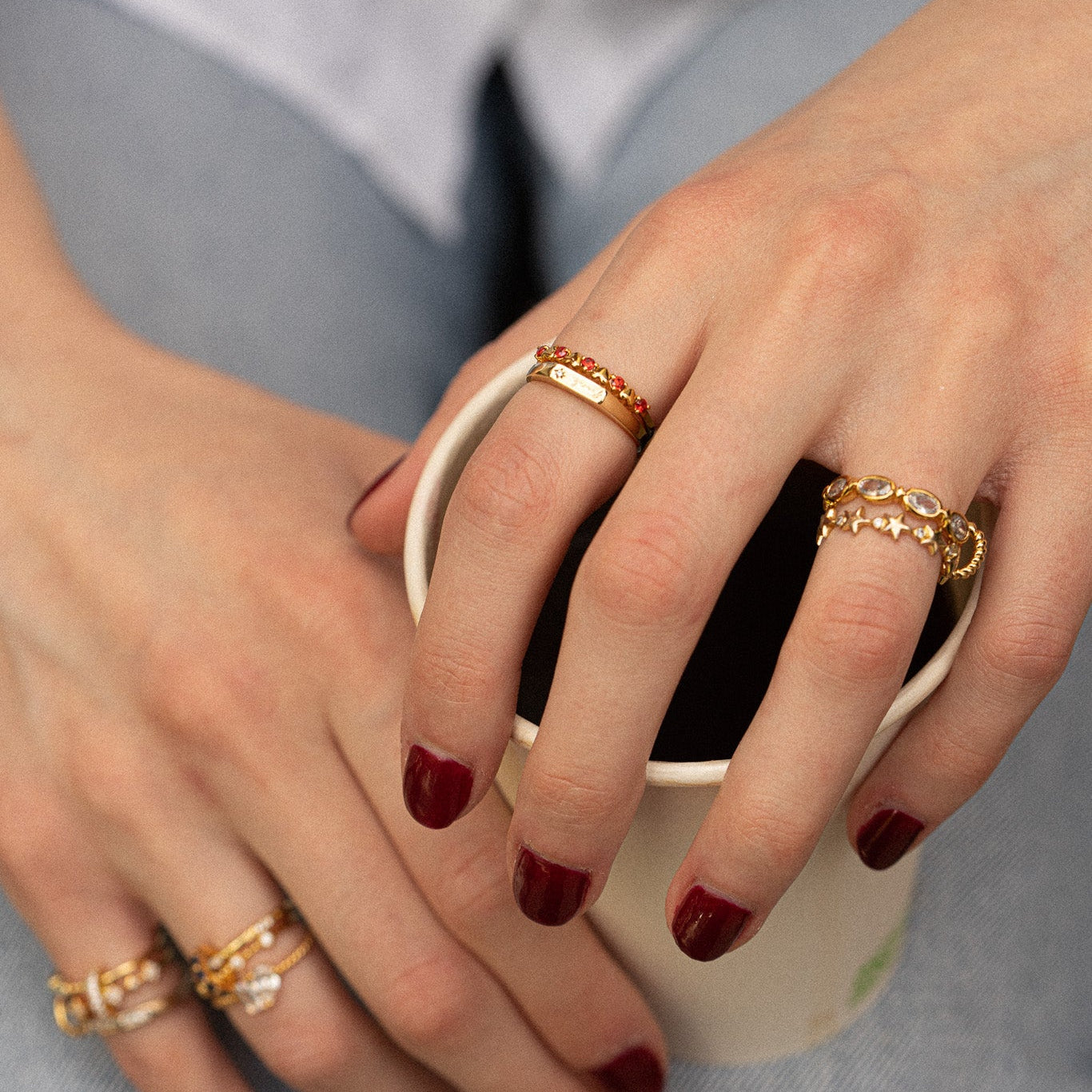 A close-up of hands with dark red nail polish wearing the Sweetheart Stacking Ring and other gold rings, holding a paper cup. The person is dressed in light blue denim jeans.
