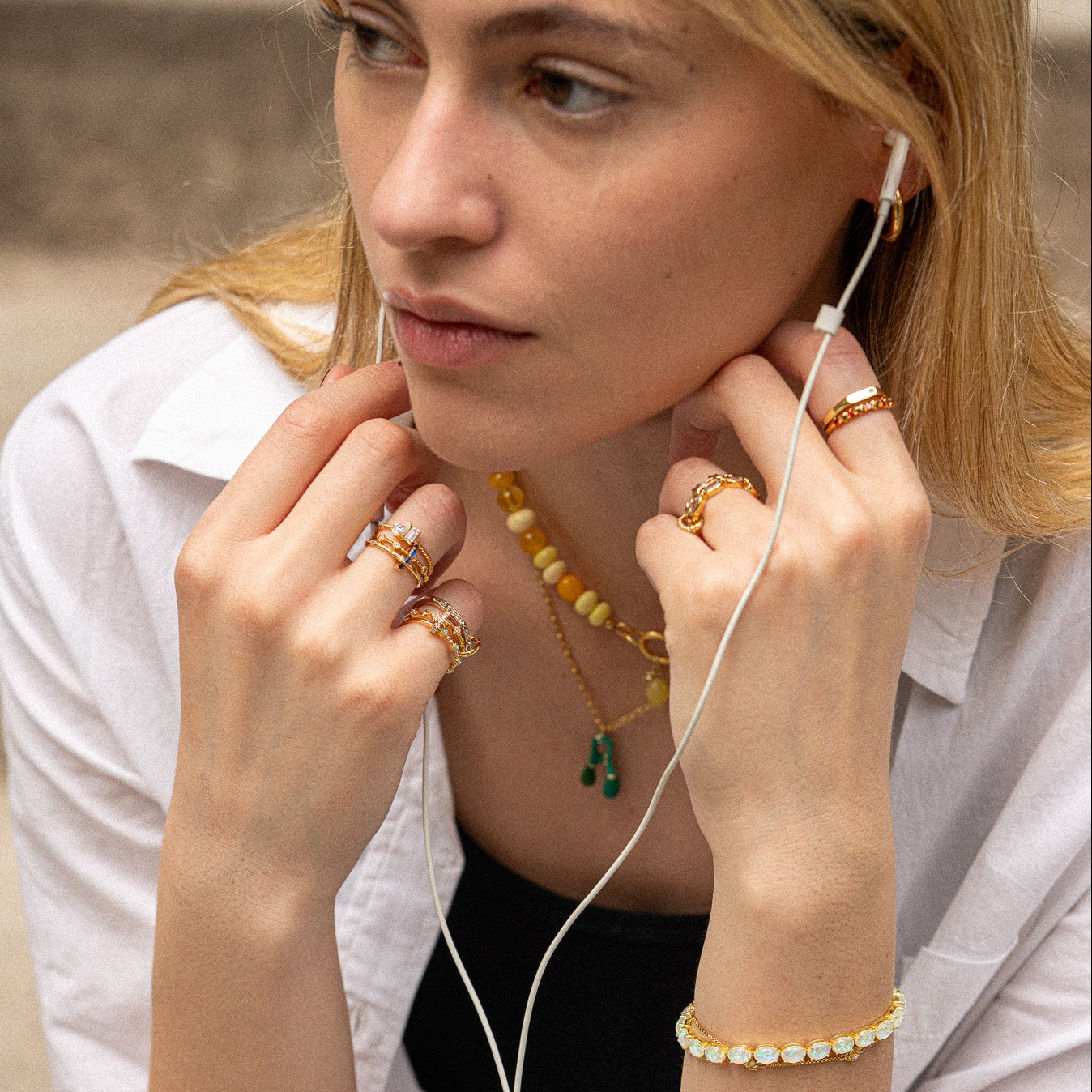 A woman with long blonde hair, wearing the Dainty Flicker Stacking Ring, a yellow beaded necklace, a bracelet, and white earphones, gazes thoughtfully to the side while touching her face with both hands.