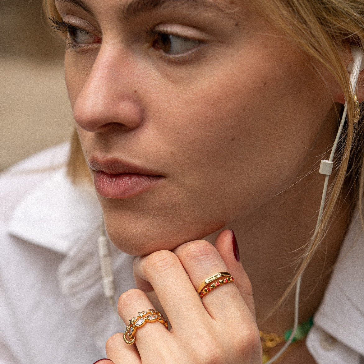 A woman with light hair wears a white shirt and earphones, her hand near her face displaying the Starlet Infinity Stacking Ring. She looks thoughtfully to the side.