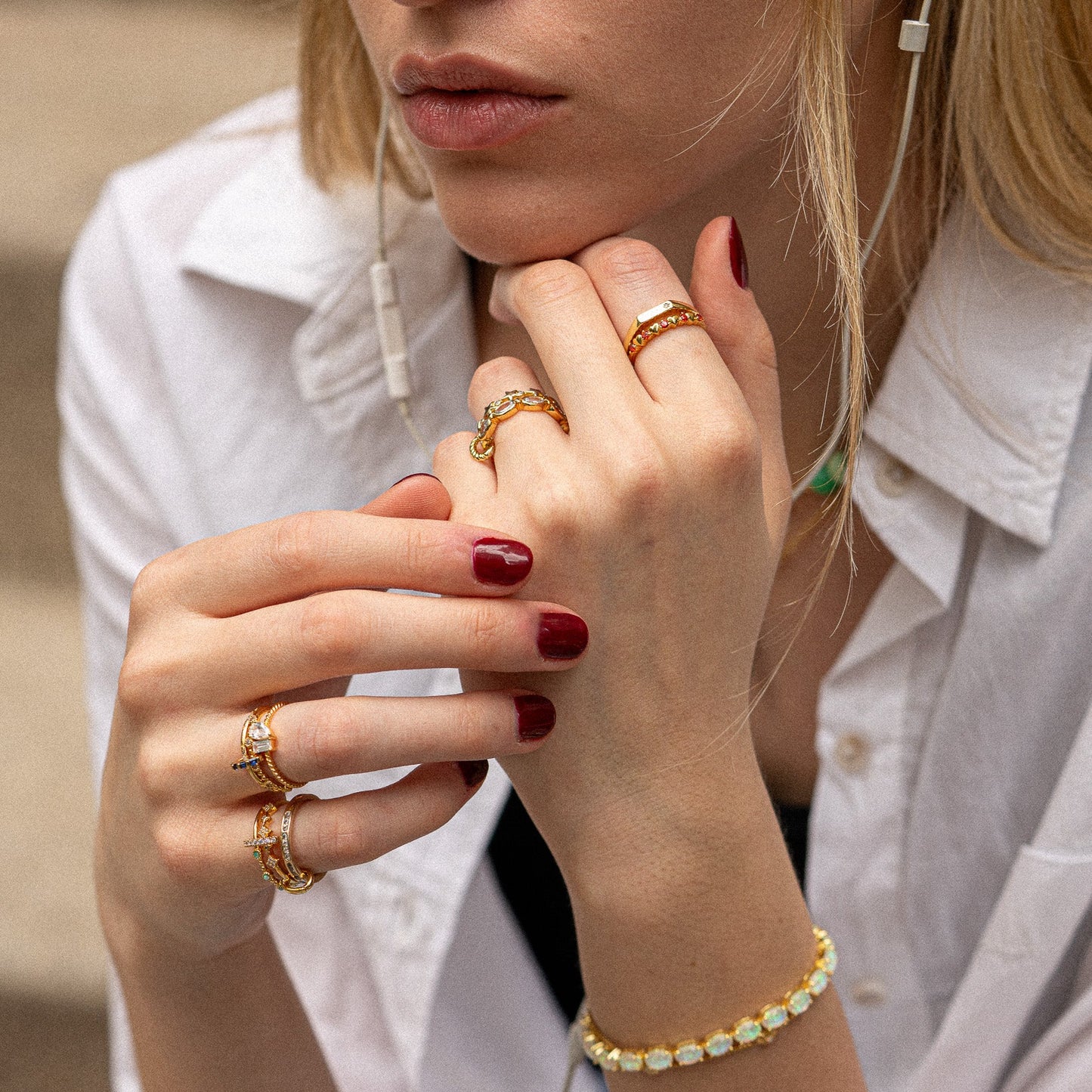 A woman with blonde hair wears the Twisted Toi Et Moi Ring and a gold bracelet, her nails painted deep red. She rests her chin on her hand, dressed in a white button-up shirt and wearing earphones.