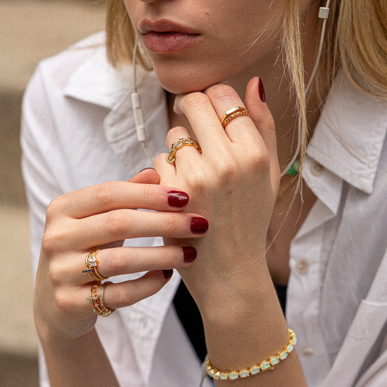 A woman with blonde hair wears the Twisted Toi Et Moi Ring and a gold bracelet, her nails painted deep red. She rests her chin on her hand, dressed in a white button-up shirt and wearing earphones.