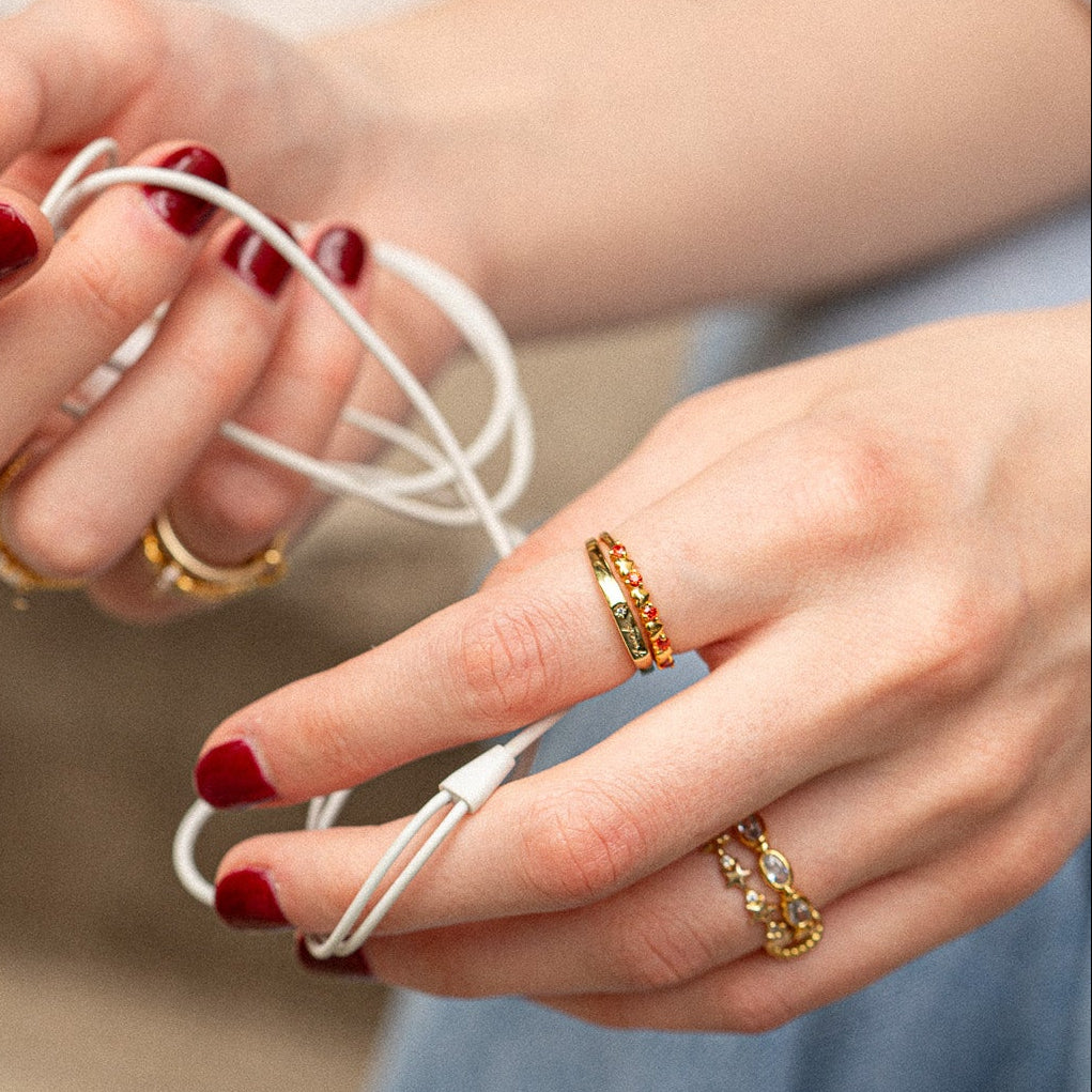 Close-up of hands holding white earbuds with red nail polish and gold rings on a blurred background